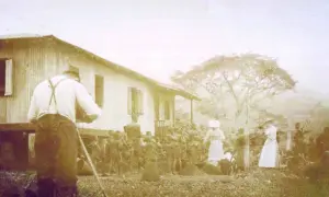 Une photo historique choquante : un homme filme une scène où des femmes en blanc semblent diriger un groupe d'indigènes devant une maison.