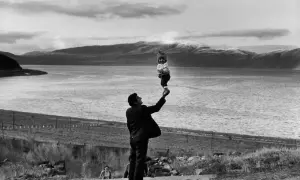Photographie artistique : un homme soulève un bébé souriant au-dessus d'un lac, avec des montagnes en arrière-plan.