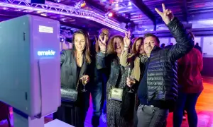 Des amis s'amusent devant un photobooth, faisant des signes de paix. L'ambiance est festive, éclairée par des lumières violettes.