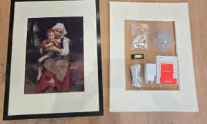 Cadre noir avec une peinture de mère et enfant, à côté d'un cadre blanc contenant des accessoires de montage, comme un niveau à bulle, un chargeur et des adhésifs, rappelant l'effet fresnel d'une œuvre d'art.
