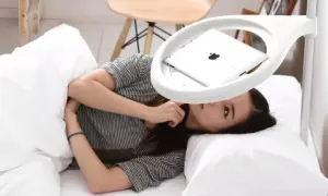 Femme allongée dans un lit, regardant une tablette posée sur un support blanc au-dessus d'elle.