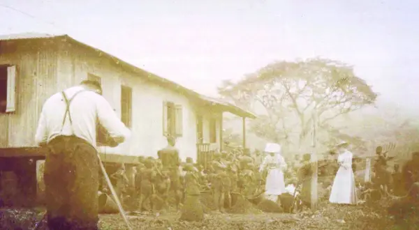 Une photo historique choquante : un homme filme une scène où des femmes en blanc semblent diriger un groupe d'indigènes devant une maison.