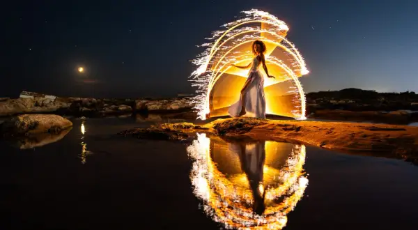 Une femme en robe blanche danse sous un ciel étoilé, entourée d'arcs de lumière incandescente. Le reflet de cette scène magique se mire dans l'eau calme, évoquant un coucher du soleil féerique.