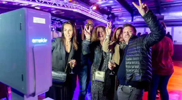 Des amis s'amusent devant un photobooth, faisant des signes de paix. L'ambiance est festive, éclairée par des lumières violettes.