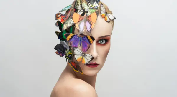 Portrait d'une femme au maquillage audacieux, ornée de papillons colorés sur la tête et le visage, évoquant une **flora borsi** onirique.
