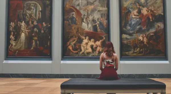 Une femme assise sur un banc, contemplant trois tableaux baroques. L'analyse photo révèle une scène de musée immersive.