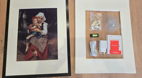 Cadre noir avec une peinture de mère et enfant, à côté d'un cadre blanc contenant des accessoires de montage, comme un niveau à bulle, un chargeur et des adhésifs, rappelant l'effet fresnel d'une œuvre d'art.