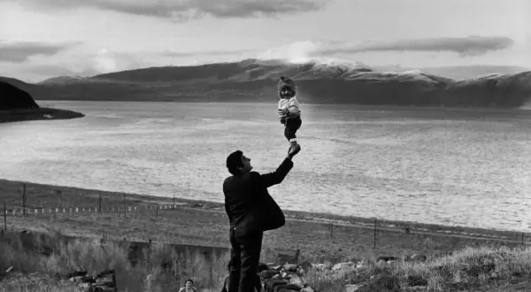 Un homme soulève un bébé au-dessus de l'eau, une scène qui semble être une mise en scène poétique avec des montagnes en arrière-plan.