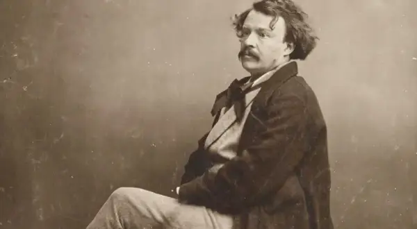 Portrait photographique 19ème siècle d'un homme à la moustache et aux cheveux bouclés, vêtu d'un manteau sombre et d'une chemise à nœud.