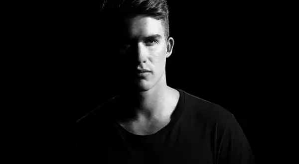 Un jeune homme au regard intense, éclairé par un côté, avec un fond noir pour mettre en valeur son visage.