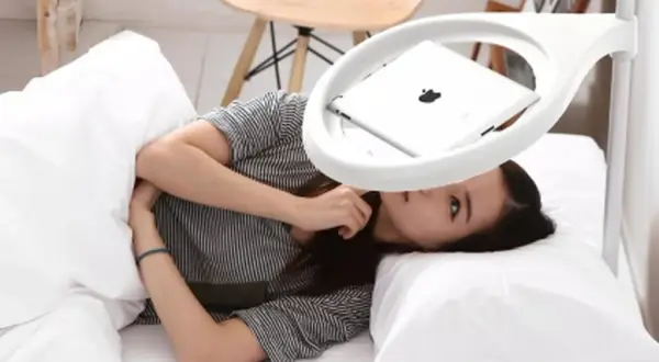 Femme allongée dans un lit, regardant une tablette posée sur un support blanc au-dessus d'elle.