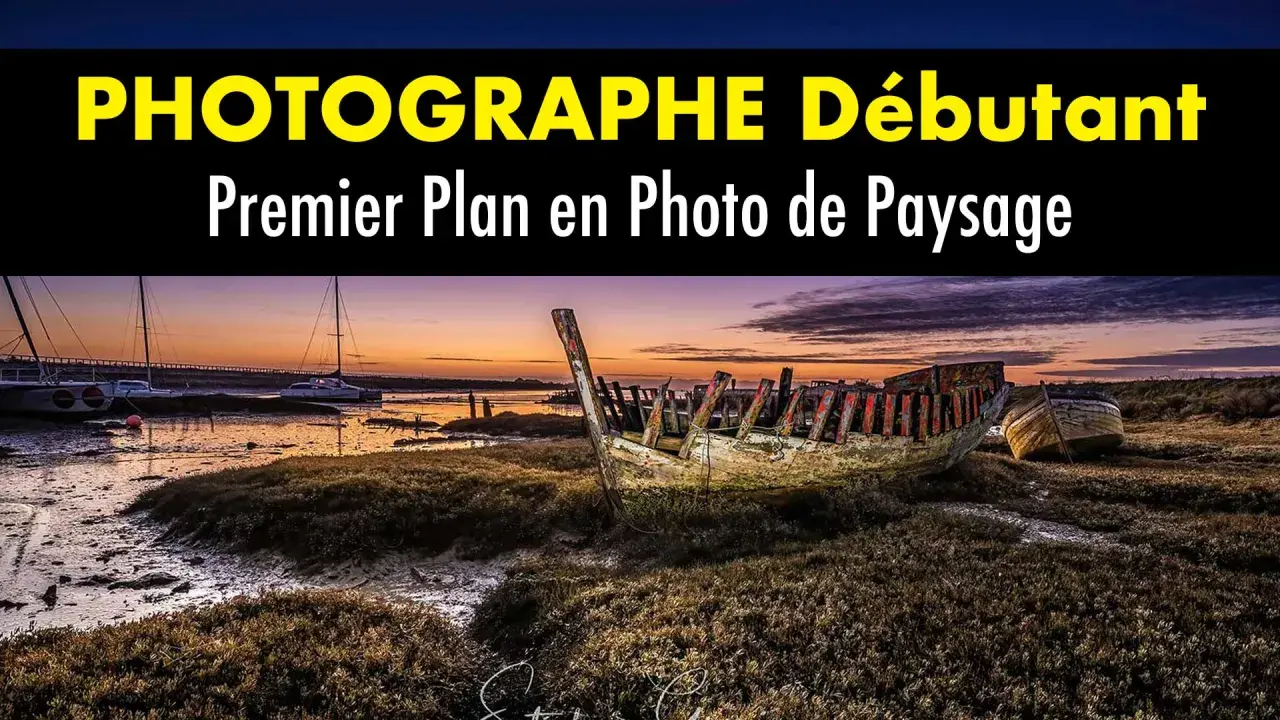 Épave de bateau en premier plan sur une plage au coucher du soleil. Au loin, des voiliers et un pont.