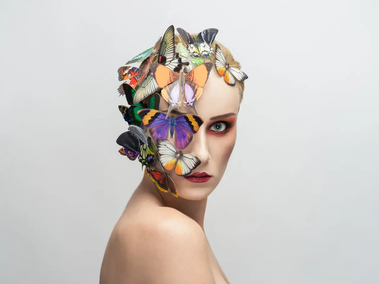 Portrait d'une femme au maquillage audacieux, ornée de papillons colorés sur la tête et le visage, évoquant une **flora borsi** onirique.