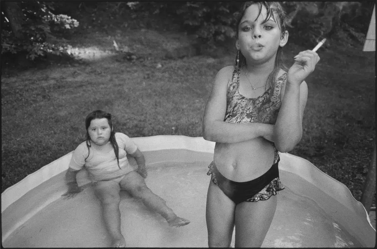 Deux jeunes filles dans une piscine gonflable, l'une fumant une cigarette, dans le style de Mary Ellen Mark.