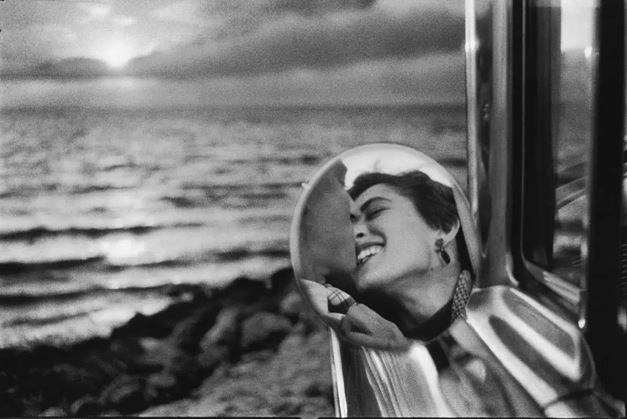 Un baiser vol&eacute;, captur&eacute; par le plus grand photographe du monde. Une femme rit, son visage refl&eacute;t&eacute; dans un r&eacute;troviseur, la mer en arri&egrave;re-plan.