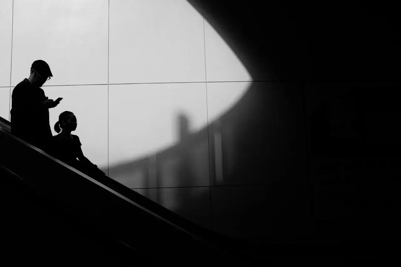 Silhouettes d'un homme et d'une femme sur un escalier mécanique, plongés dans l'ombre. La technologie de l'image noir et blanc accentue le contraste.