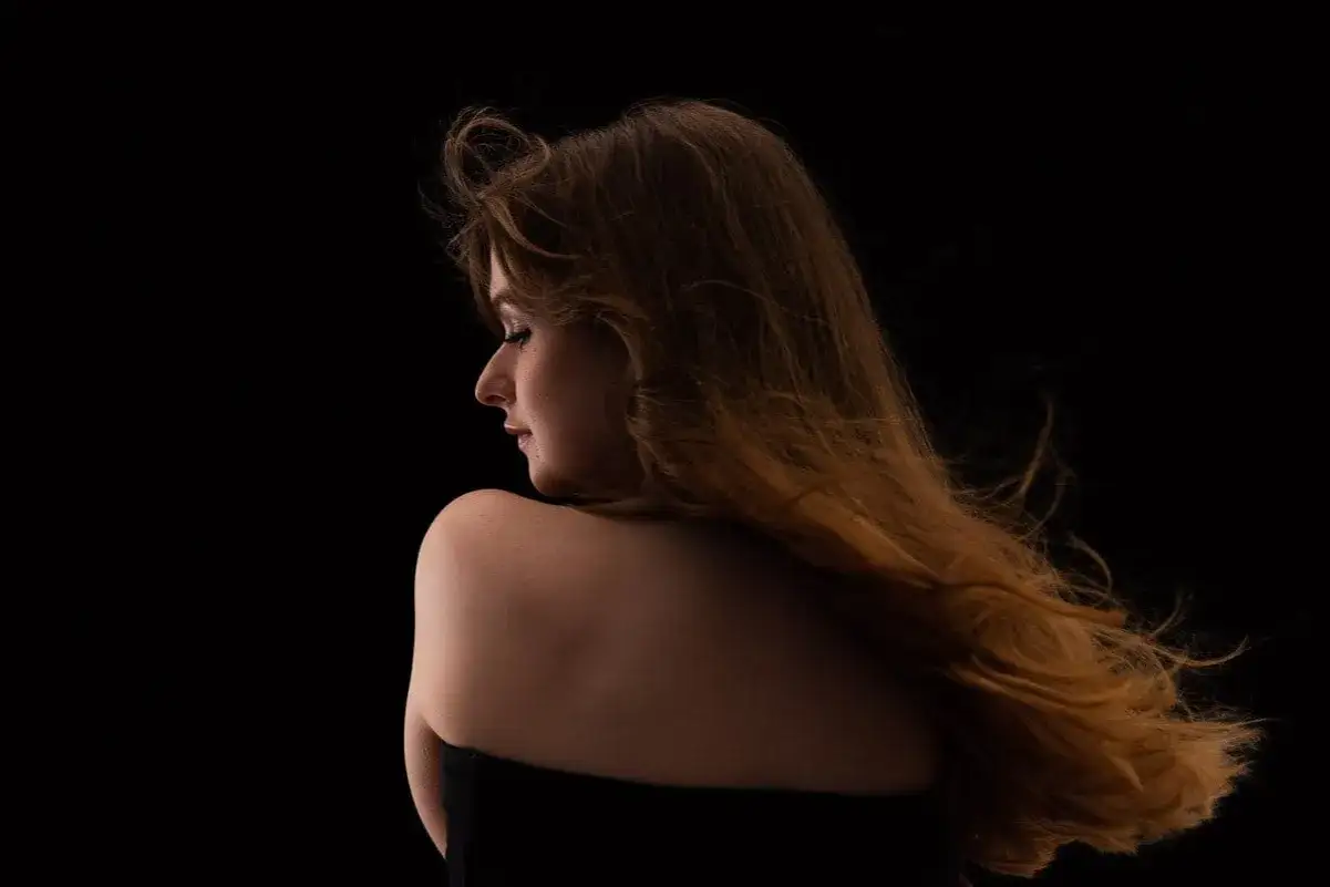 Une jeune femme aux cheveux roux longs et ondulés, vue de profil, se détache sur un fond noir. L'effet de mettre un fond noir sur une photo accentue la douceur de sa peau et la texture de ses cheveux.