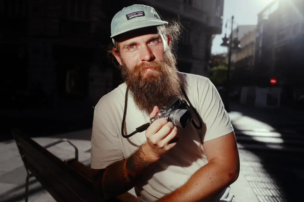 Homme barbu avec casquette Patagonia tenant un appareil photo Leica Q3 pour un test. Lumi&egrave;re du soleil sur son visage.
