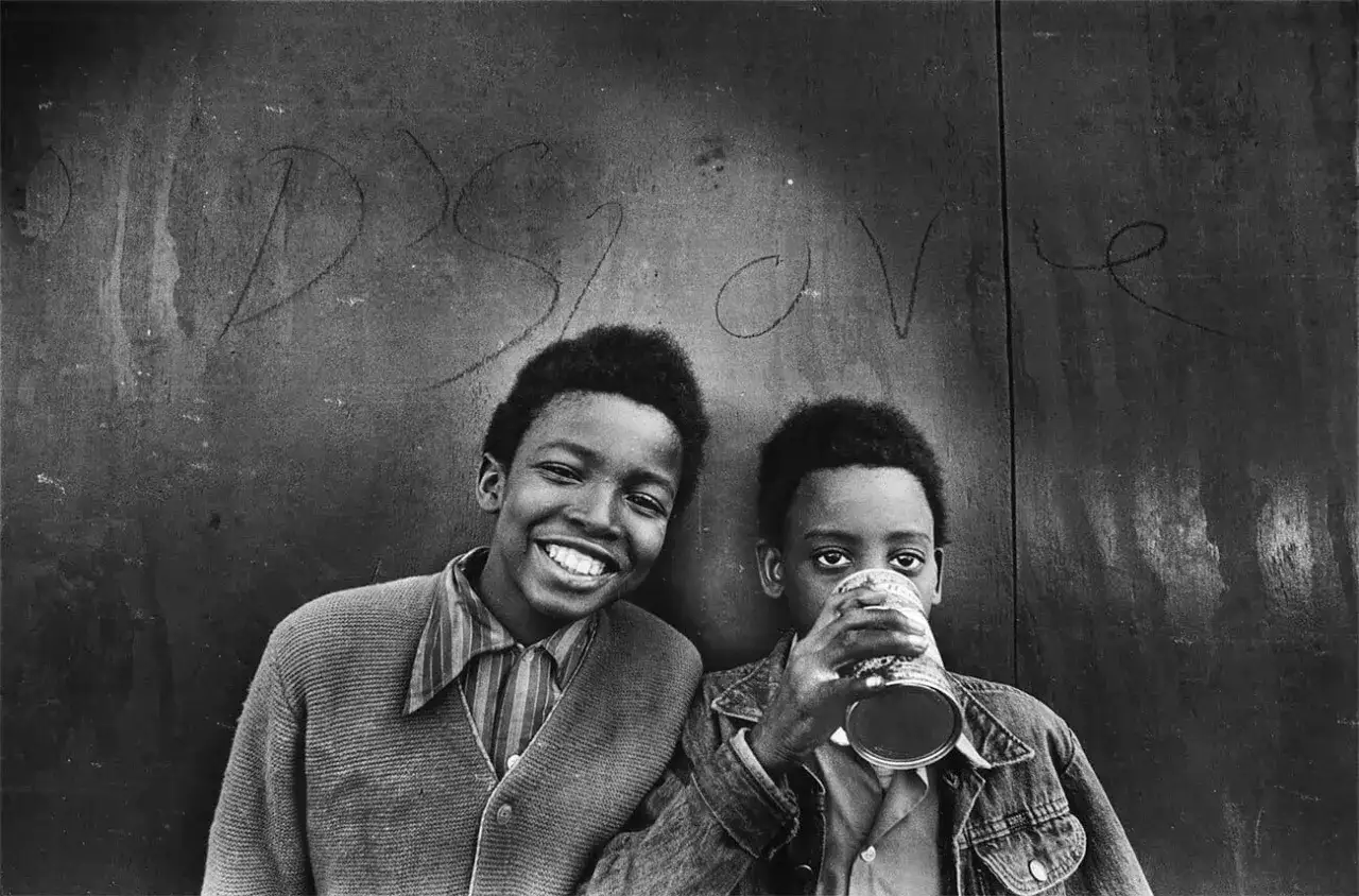Deux jeunes garçons devant un mur couvert de graffitis. L'un sourit, l'autre boit dans une canette. Une photographe femme américaine a capturé ce moment.