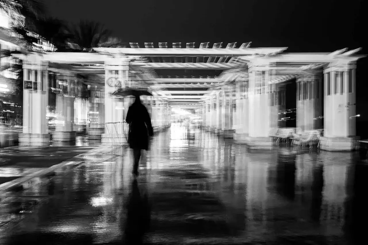 Une silhouette solitaire sous un parapluie traverse une allée de photo de rue mouillée et réfléchissante, sous une structure architecturale floue.