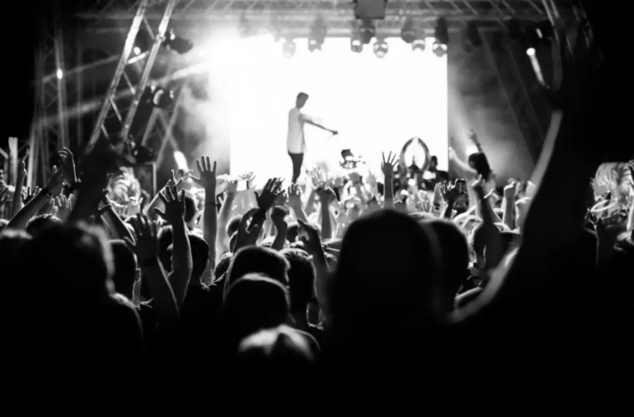 Une foule en d&eacute;lire l&egrave;ve les mains lors d'un photo concert. La sc&egrave;ne est &eacute;clair&eacute;e par des projecteurs, un artiste est visible au centre.