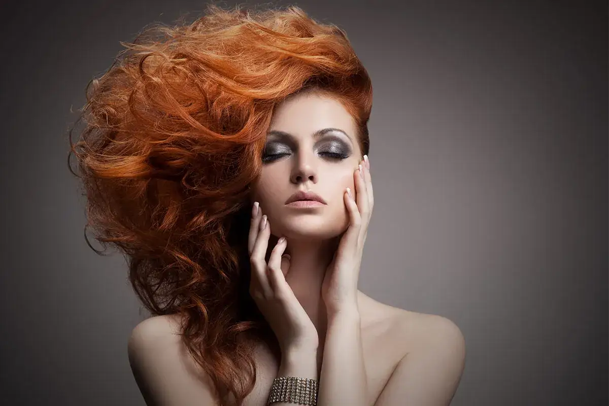 Femme aux cheveux roux flamboyants, maquillage intense, mains sur le visage. Le vignettage photo accentue le myst&egrave;re.