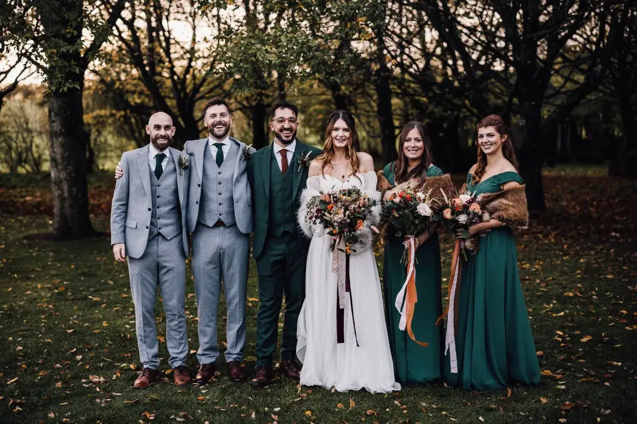 Un groupe joyeux pose pour des photos de mariage en extérieur, entouré d'arbres automnaux. La mariée, rayonnante, tient un bouquet coloré.