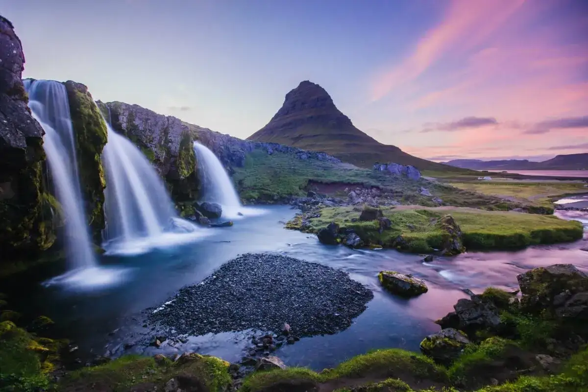 Cascades islandaises capturées en pose longue, avec le mont Kirkjufell en arrière-plan sous un ciel aux teintes roses et bleues.