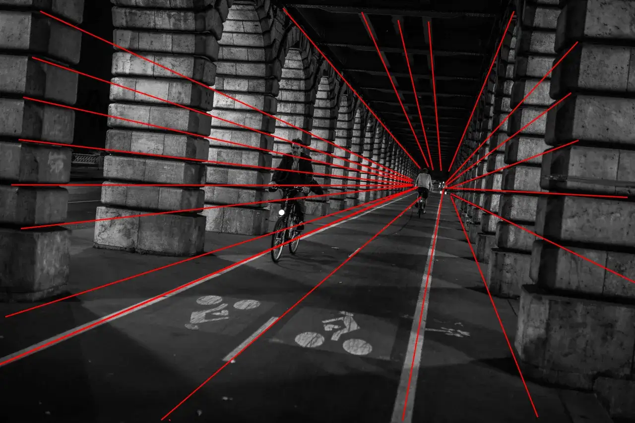 Sous un pont, deux cyclistes avancent dans un tunnel de pierre. La perspective est accentuée par des lignes rouges.