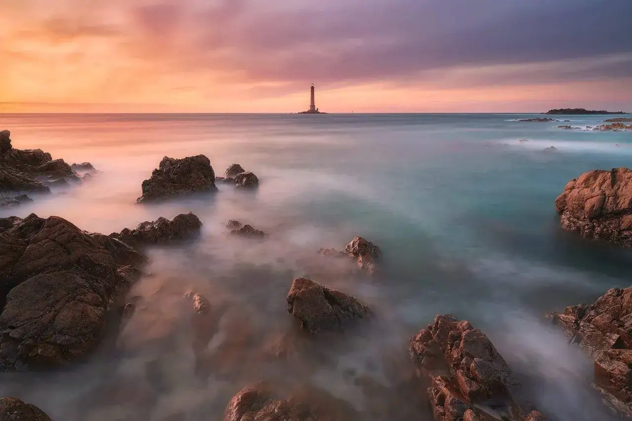 Un phare se dresse au loin sur une île rocheuse, baigné par les couleurs douces d'un coucher de soleil. L'eau, rendue soyeuse par un filtre ND, caresse les rochers au premier plan.