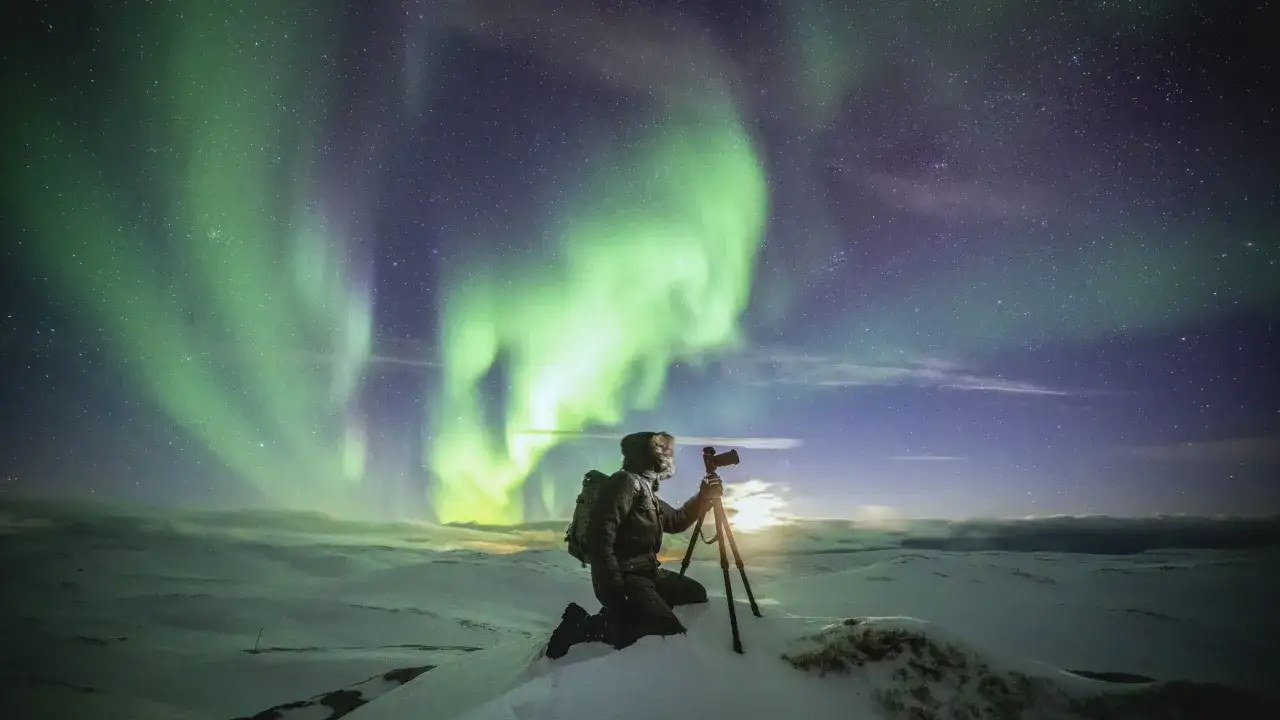 Un photographe, arm&eacute; de son tr&eacute;pied, capture la magie des aurores bor&eacute;ales vertes et violettes dans un paysage enneig&eacute;.