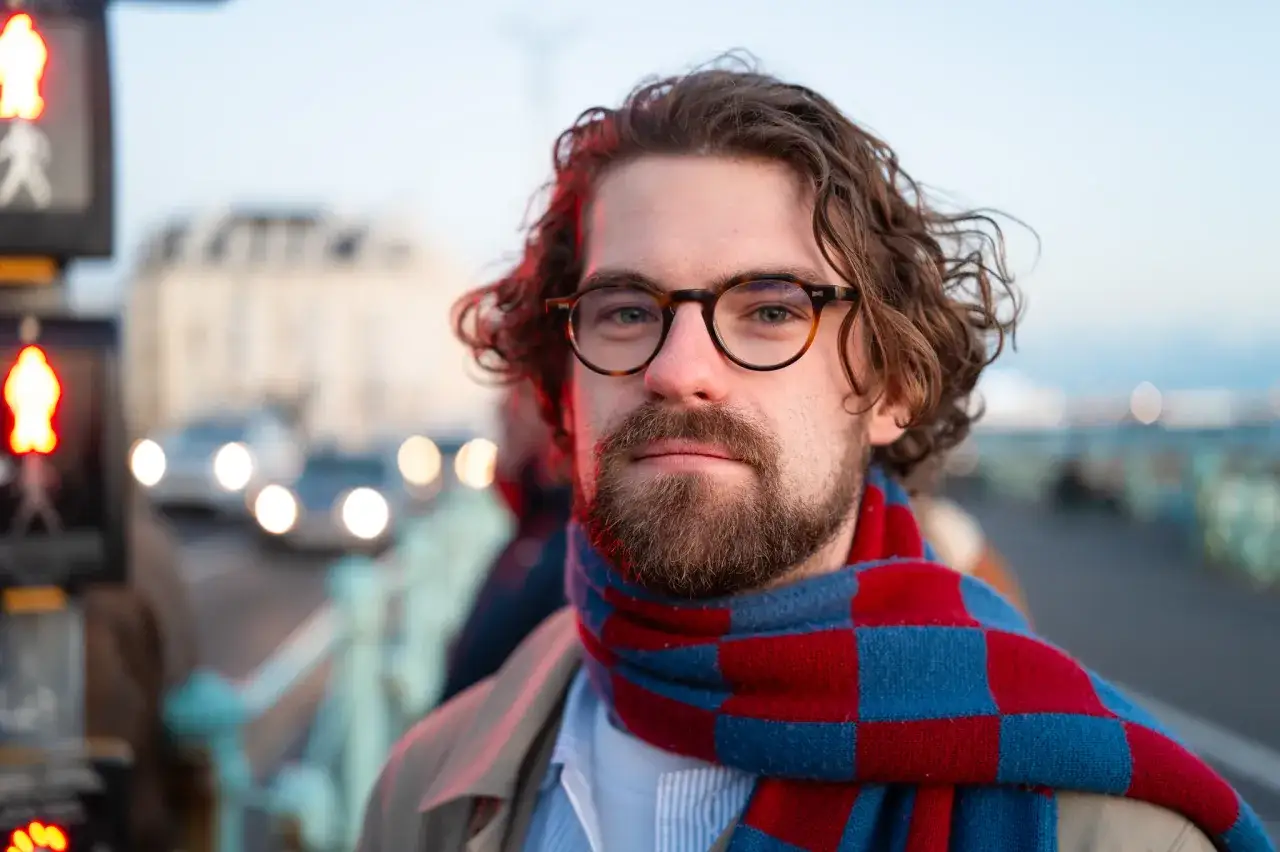 Un homme avec des lunettes et une &eacute;charpe &agrave; carreaux rouges et bleus, captur&eacute; lors d'un test du Leica Q3 dans un environnement urbain.