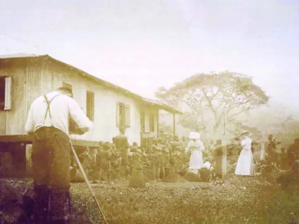 Une photo historique choquante : un homme filme une scène où des femmes en blanc semblent diriger un groupe d'indigènes devant une maison.
