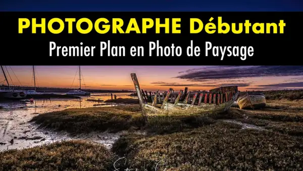 Épave de bateau en premier plan sur une plage au coucher du soleil. Au loin, des voiliers et un pont.