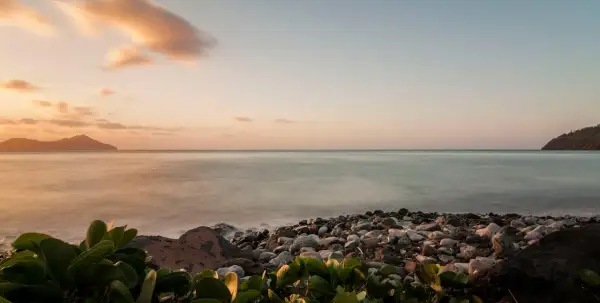 Coucher de soleil sur une plage de galets, avec des îles au loin. Les **filtres photo** accentuent les tons pastel du ciel et la douceur de l'eau.