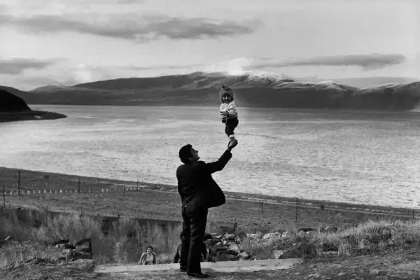 Photographie artistique : un homme soulève un bébé souriant au-dessus d'un lac, avec des montagnes en arrière-plan.