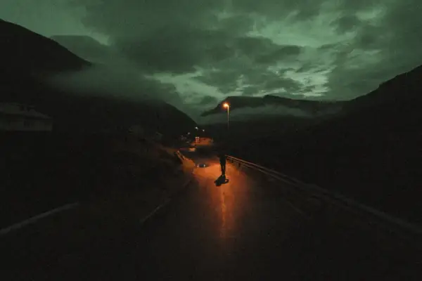 Une silhouette solitaire sur une route de montagne, éclairée par un lampadaire. Le ciel est sombre et nuageux, donnant à cette **grain photo en ligne** une atmosphère mystérieuse.