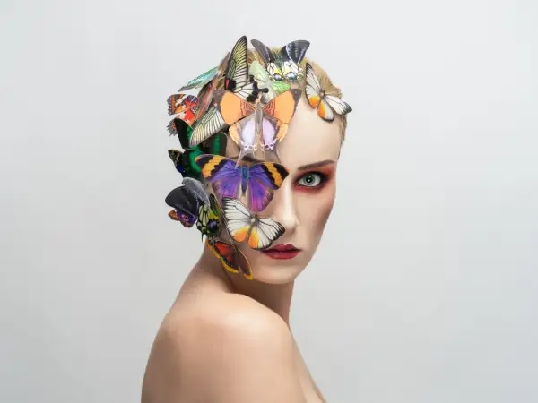 Portrait d'une femme au maquillage audacieux, ornée de papillons colorés sur la tête et le visage, évoquant une **flora borsi** onirique.