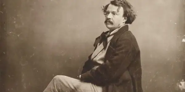 Portrait photographique 19ème siècle d'un homme à la moustache et aux cheveux bouclés, vêtu d'un manteau sombre et d'une chemise à nœud.