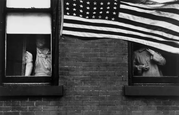 Une photographe femme américaine capture deux femmes regardant par des fenêtres, un drapeau américain flottant au-dessus.